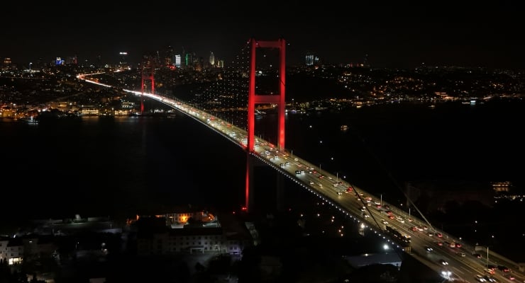 Aerial night view of a red suspension bridge spanning a dark body of water with a city skyline lit up in the background, and traffic along the bridge