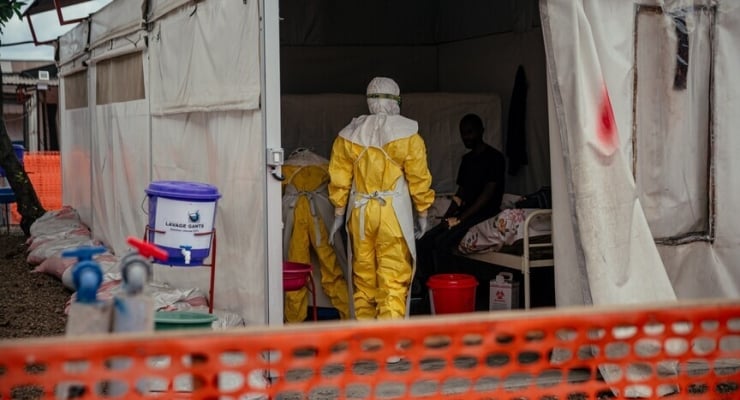 Healthcare workers in yellow hazmat suits tending a patient inside a white medical tent; orange safety barrier in the foreground.