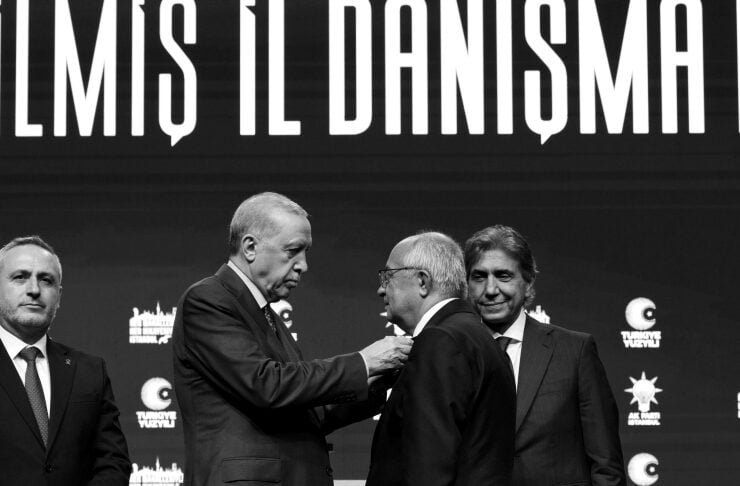 Four men in business suits on a stage during a formal ceremony; one man pins an item on another as a third watches, with a large banner and logos in the background (black-and-white photo).