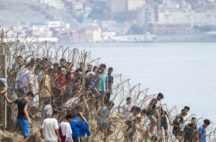 People walk along a barbed-wire fence on a rocky slope by the sea, with a city in the background.