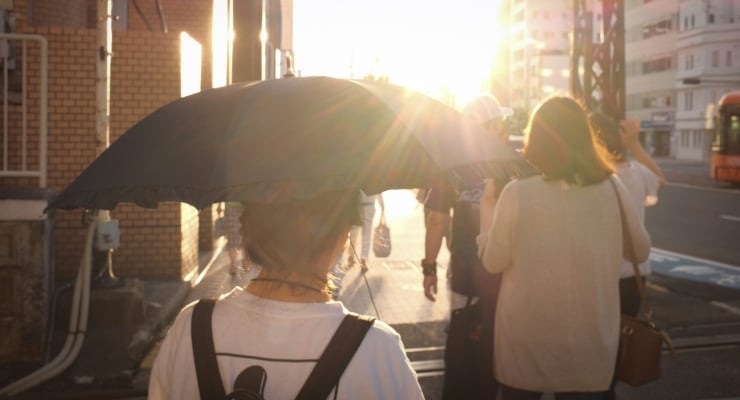 People walk along a sunlit city sidewalk; a person with a backpack holds a large umbrella shielding others from the bright sun.