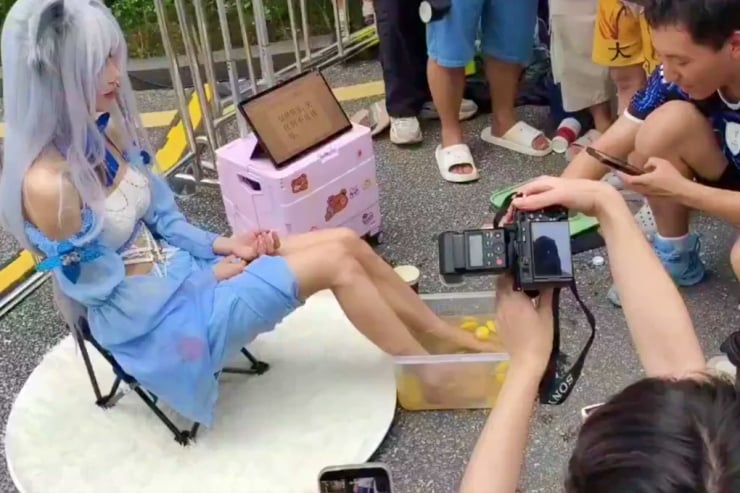 Cosplayer in a light blue outfit and long white wig sits on a chair as photographers film and take pictures nearby outdoors on a street.