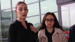 Two women stand indoors by a glass wall, looking serious; one in a black top with a ponytail, the other in a beige coat and glasses.