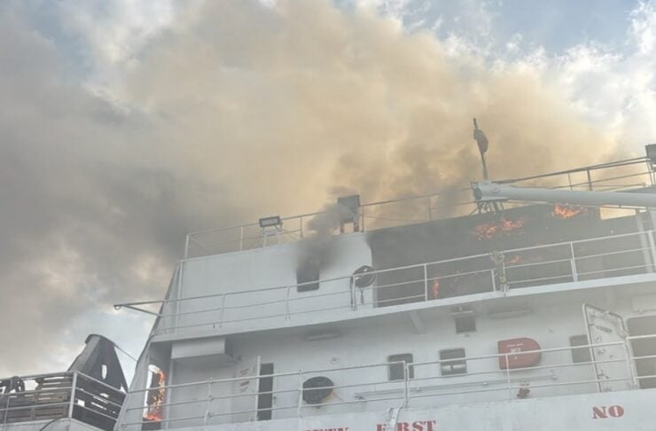 Fire on the upper deck of a ship with dark smoke billowing from flames near railings; safety signage reads 'Safety First' on the hull.
