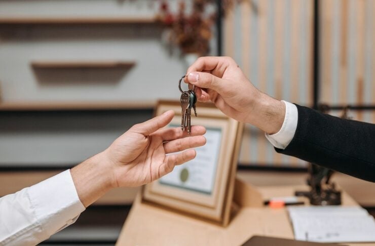 Two hands exchange a set of keys across a desk in an office setup, suggesting a handover or handoff of a property or vehicle key.