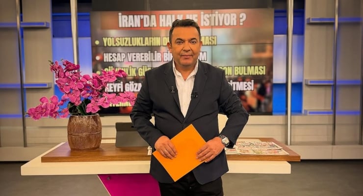 Male news anchor in a TV studio, holding an orange envelope, standing at a desk with a pink orchid and a large screen behind displaying Turkish text.