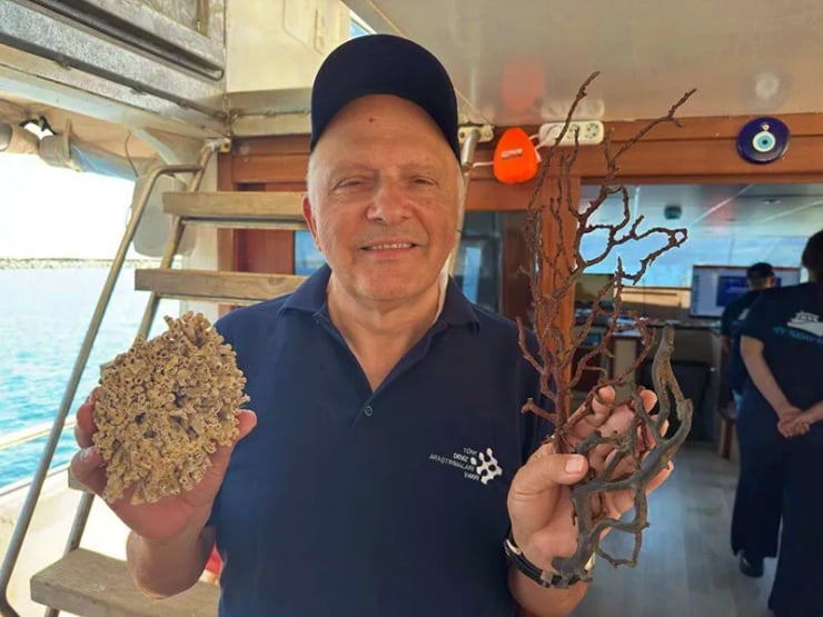 Smiling man on a boat holding brain coral in one hand and branching coral in the other, with the ocean in the background.