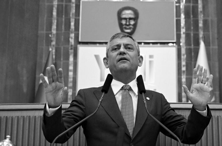 Politician in a suit delivers a speech at a podium with two microphones, hands raised, banner reading 'YENİ PARTİ' in the foreground.