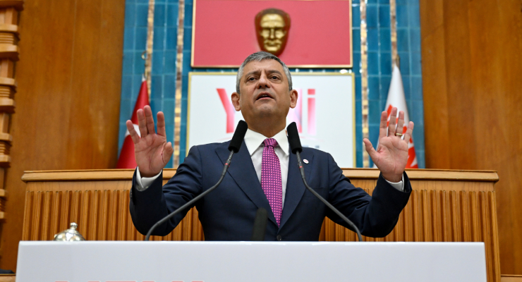 Man in a dark suit and pink tie speaks at a podium with two microphones, hands raised, in a formal government chamber with flags and a gold bust in the background.