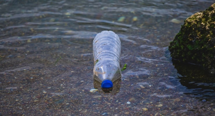 Plastic bottle with blue cap partially submerged in shallow water near a rocky shoreline.