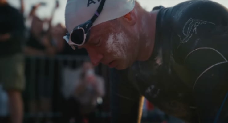 Triathlete wearing a swim cap and goggles bends over, exhausted, with a cheering crowd in the background.