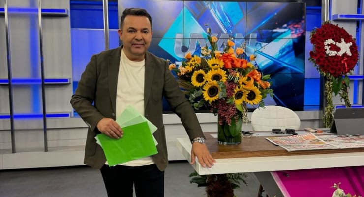 Male TV host in a studio, standing by a desk with a large bouquet of sunflowers and other flowers, holding green papers, with a news set backdrop behind him.