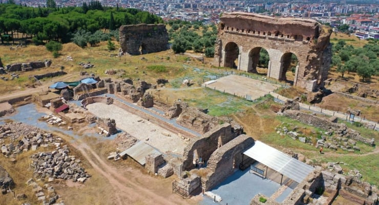 Ancient ruins with large arched stone structure on a hill and a distant city skyline in the background.