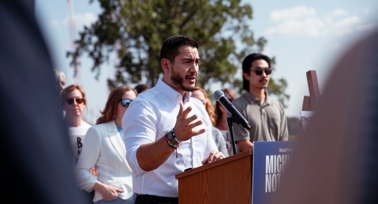 Man in white shirt speaking at a wooden podium with a microphone, gesturing with his hand during an outdoor rally or press event, supporters in the background; sunny day