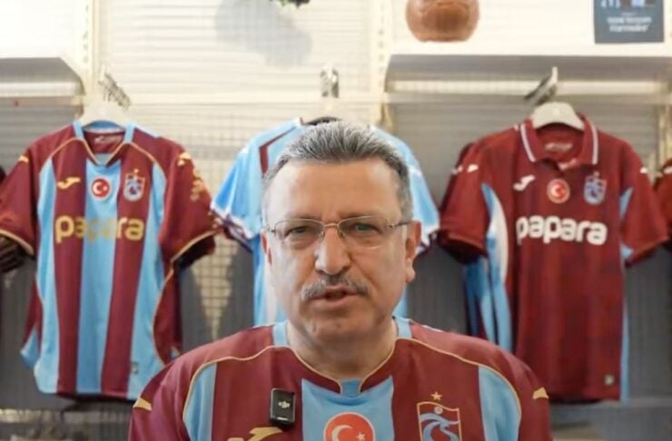 Man wearing a maroon and light-blue striped football jersey stands in a shop with matching jerseys on racks behind him, including a visible Papara sponsor logo and a Turkish flag crest on the chest.