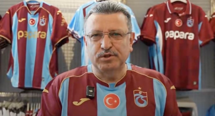 Man with glasses wearing maroon and light-blue striped football jersey, speaking to camera with club jerseys hanging behind him in a studio/store.
