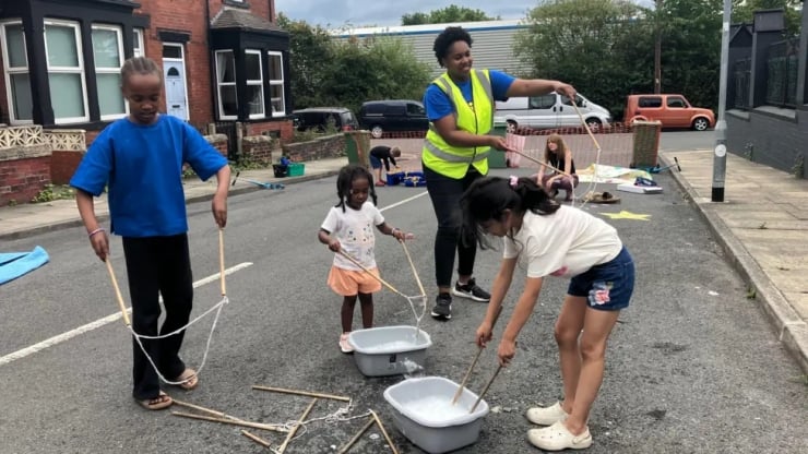Group of children on a street, supervised by a woman in a high-visibility vest, weaving with sticks and strings over two buckets in water.