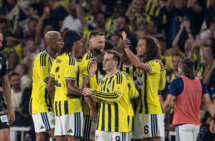 A group of footballers in yellow jerseys celebrate on the field, high-fiving and embracing a teammate amid cheering fans.