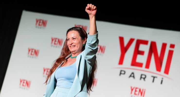 Woman on stage with fist raised in triumph, backdrop reads YENİ PARTİ banners.