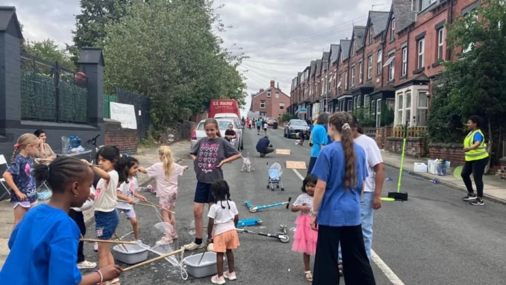 A sunny urban street blocked off for a kids' event with children and volunteers playing with toy scooters and buckets along the curb.