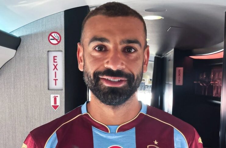 Portrait of a man with a beard wearing a maroon and light-blue striped soccer jersey with a Turkish flag crest, standing indoors near an exit sign.