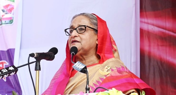 A woman in a red and gold sari speaking at a podium with two microphones and a backdrop behind her.