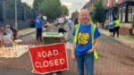 Smiling volunteer in a blue t-shirt and high-visibility vest stands at a road closure with a red 'ROAD CLOSED' sign; families and kids in the background at a community event.