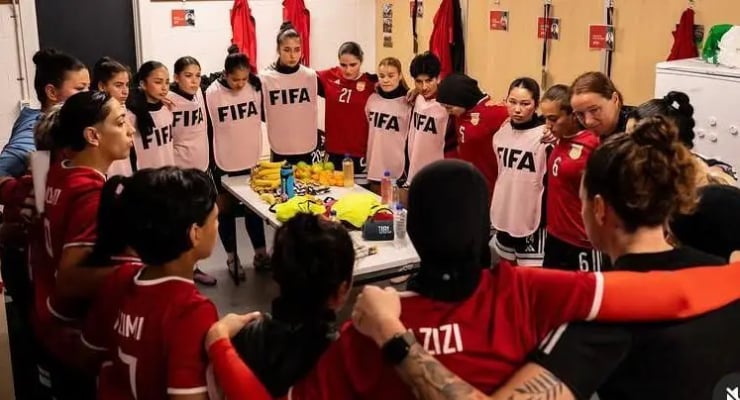 Women’s soccer players in red uniforms form a huddle in a locker room around a table with water bottles and gear, wearing light pink FIFA bibs for training or warmups.