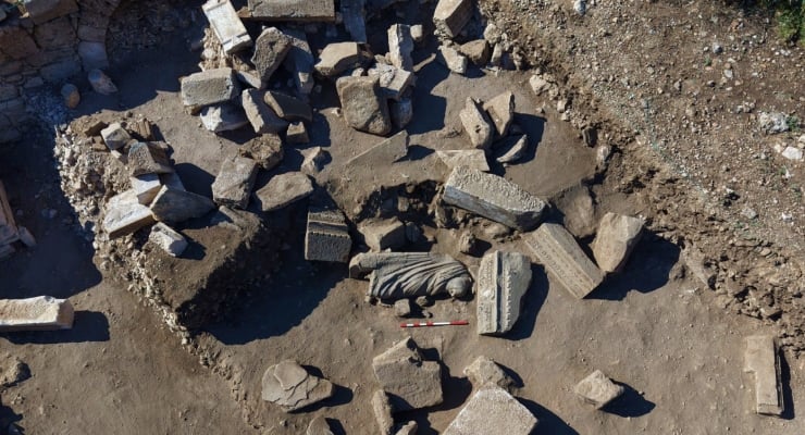 Archaeological dig with scattered stone blocks and a carved hand fragment, plus a measurement scale nearby.