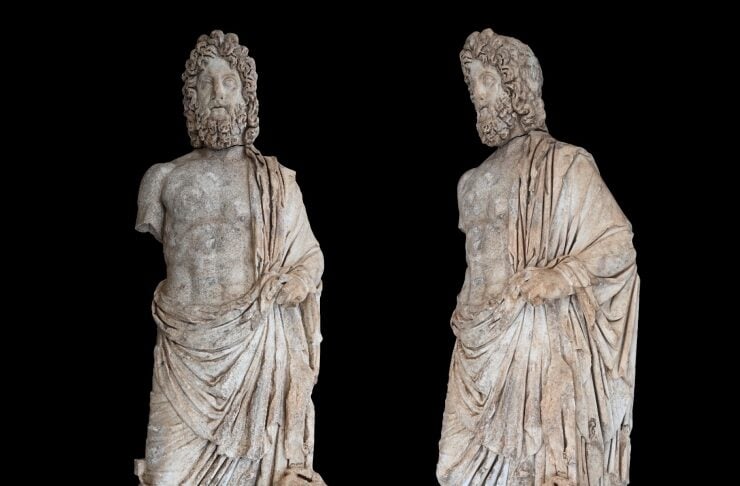 Two ancient stone statues of bearded men in draped robes standing on bases against a black background, with a measurement scale at the bottom.