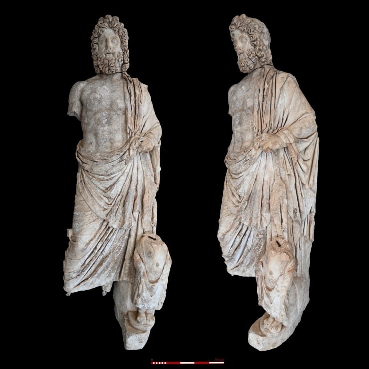 Two ancient stone statues of bearded men in draped robes standing on bases against a black background, with a measurement scale at the bottom.