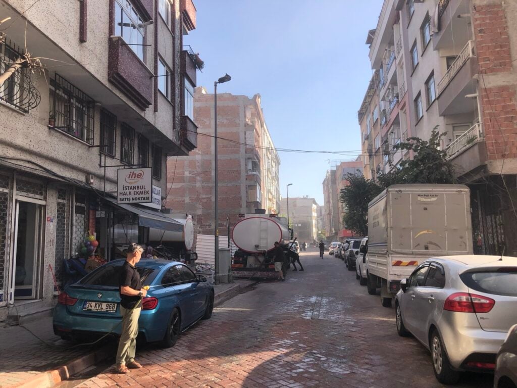 Narrow street in a residential area; a man waters a blue car while a large tanker truck sits mid‑street.