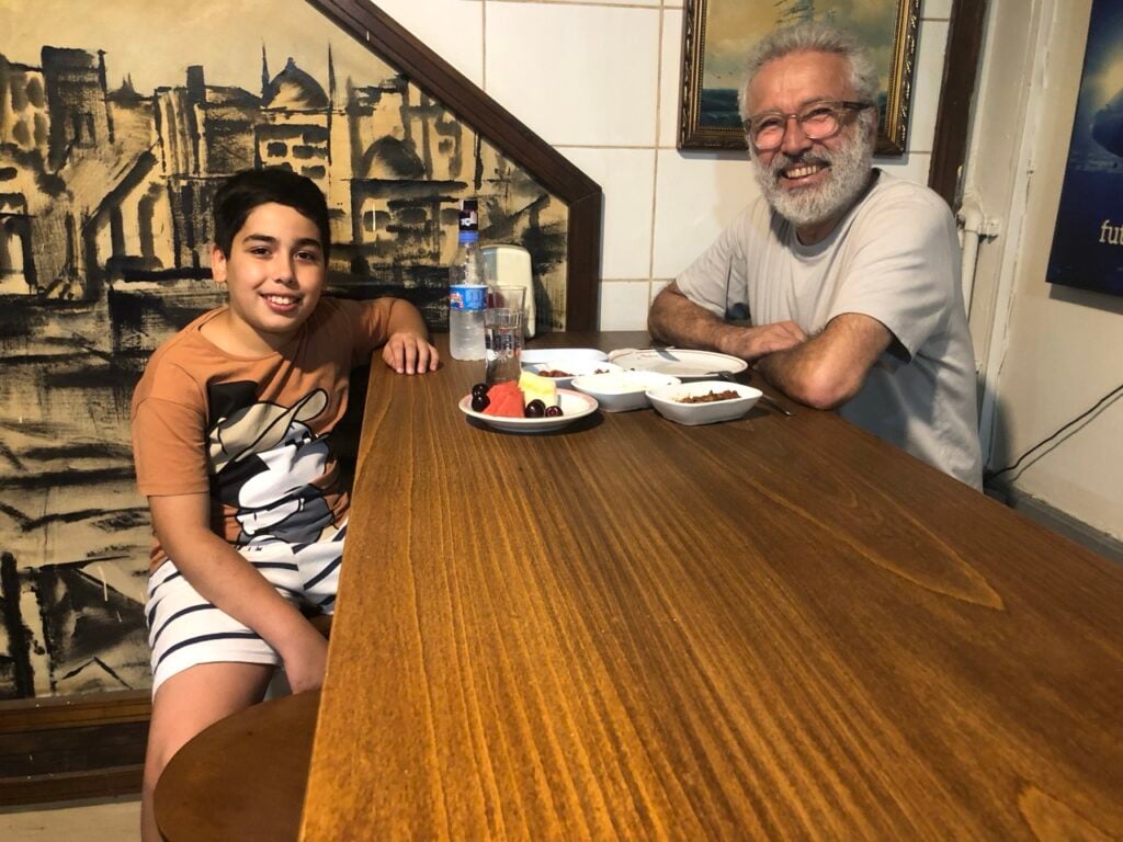Boy and older man (grandfather) smile at the camera while seated at a wooden dining table in a restaurant, with plates of food and a bottle of water between them.