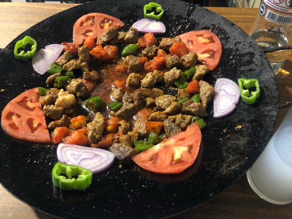 Diced beef with green bell peppers and tomatoes on a round black plate, garnished with tomato wedges and red onion slices.