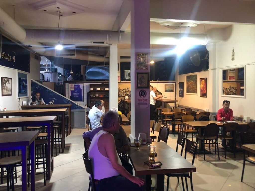 Cafe interior with high tables, stools, and a purple central pillar, framed art on the walls, and patrons seated and talking.