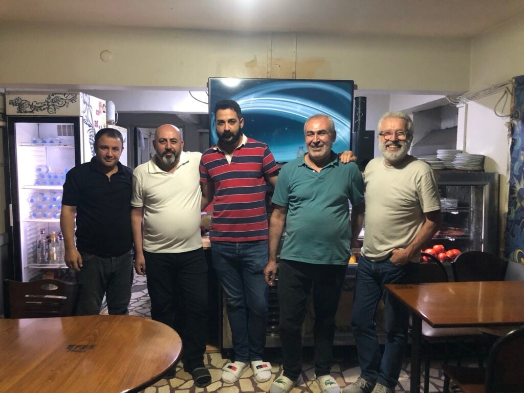 Five men pose together in a casual dining room, with a fridge and fruit display behind them.