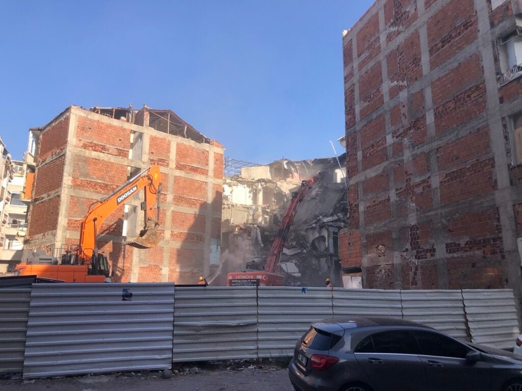 Construction site with an orange Doosan excavator demolishing a brick building, dust rising between rubble behind a corrugated fence.