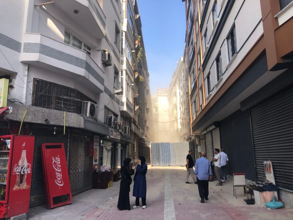 Narrow urban alley with tall residential buildings, shuttered shops, and a dust-filled construction barrier at the far end; several pedestrians nearby.
