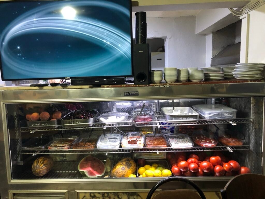 Cafe display fridge with assorted fruits, desserts, and plates on top; a TV and speaker system sit above.