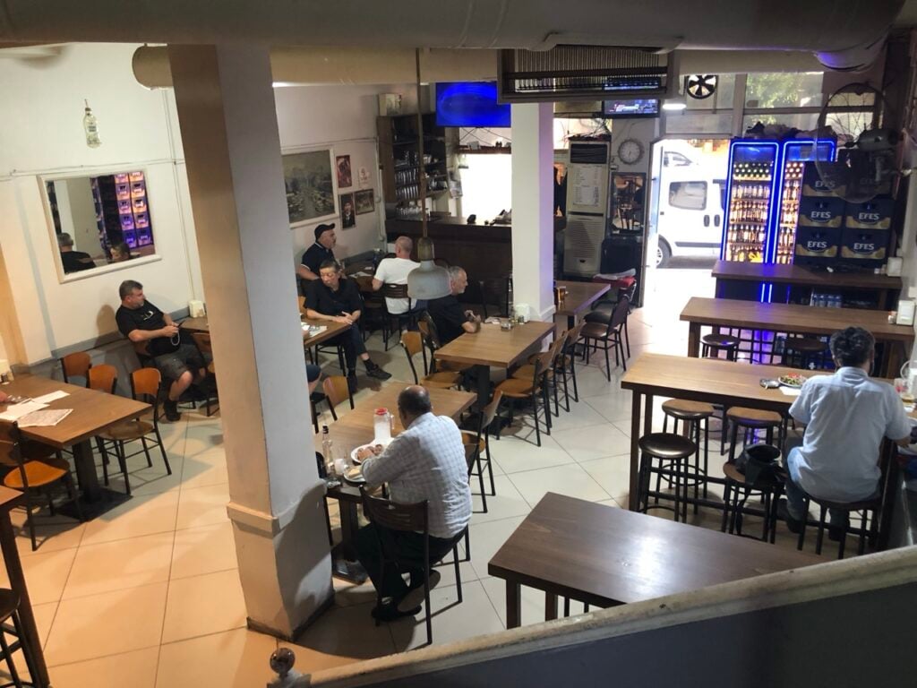 Small cafe or tavern interior with several patrons seated at wooden tables and stools, pillar in the center and a bar area in the background.