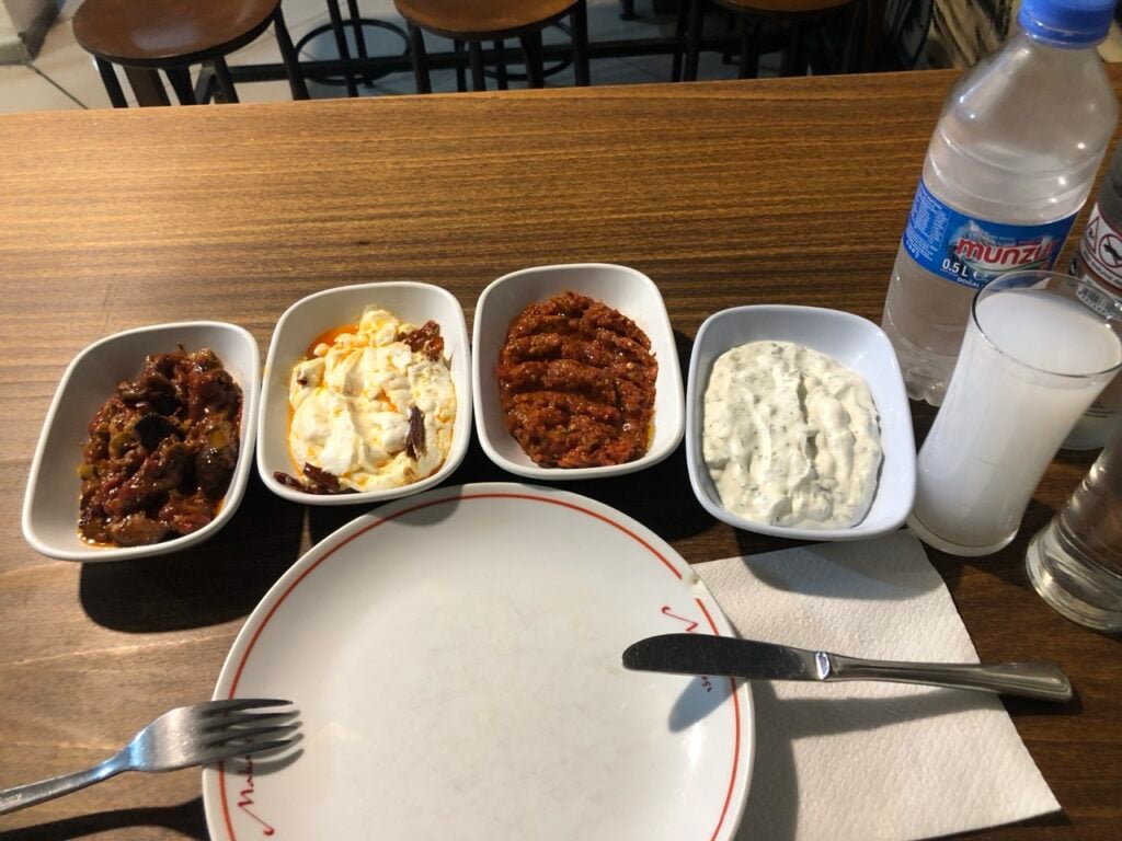 Four white bowls with different foods—meat stew, eggs with peppers, spiced meat strips, and a creamy yogurt dip—on a wooden table with a water bottle and glass in view.