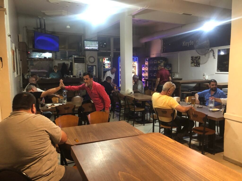 Inside a casual bar/restaurant with wooden tables and diners; a man in a red shirt and another man share a high-five across a table.