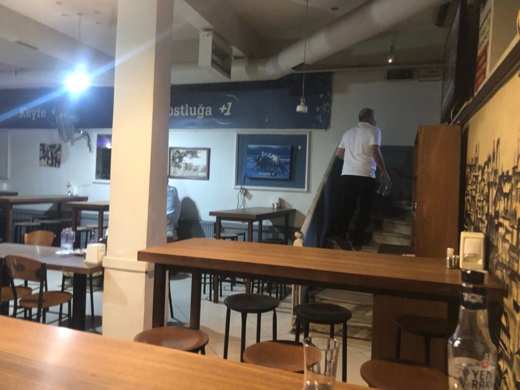 Inside a casual cafe with wooden tables and stools; a man in a white shirt walks up a staircase in the back near a blue wall sign.