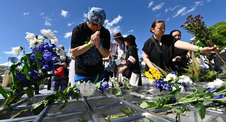 People lay colorful flowers on a memorial rack during an outdoor remembrance ceremony under a bright blue sky.