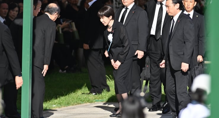 People in black suits bowing in a formal outdoor ceremony; a woman with a white corsage bows among dark-clad attendees.