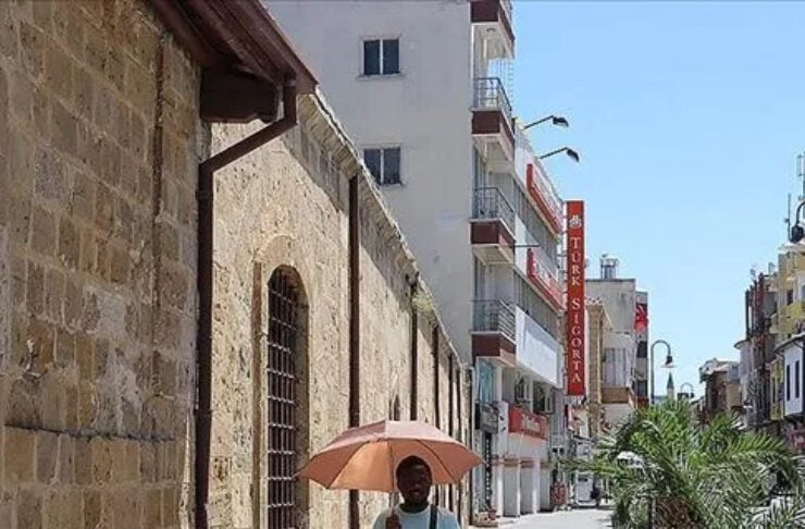 A man walks down a sunny street with a peach umbrella, beside a stone wall and palm trees lining the sidewalk.