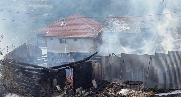 Burned wooden shed with charred walls and rising smoke, with a two-story house and red-tiled roof in the background amid a rural fire scene.