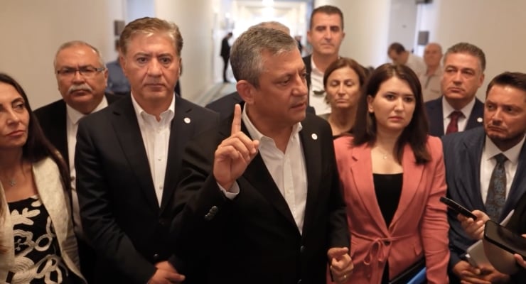 Man in a dark suit raises his index finger while speaking to a group of officials in a hallway; reporters and colleagues surround him.