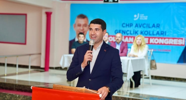 Man in a suit speaking into a microphone at a podium during a formal event, with a blue banner and panelists in the background.