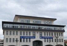 White city hall building with arched ground-floor entrances and blue Kusadasi Belediyesi signs, set on a grassy lawn in front.